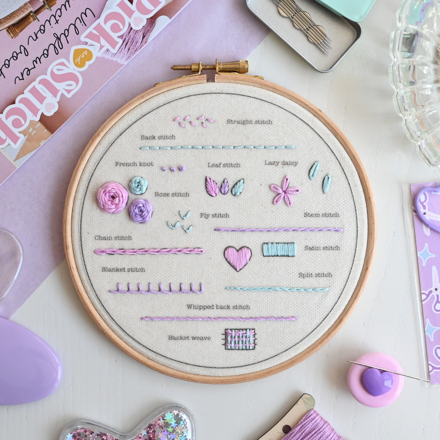 A wooden embroidery hoop displaying various stitch patterns and names on natural fabric, surrounded by craft supplies including a spool of lavender thread, a pin cushion, and a booklet on a pastel desk background.