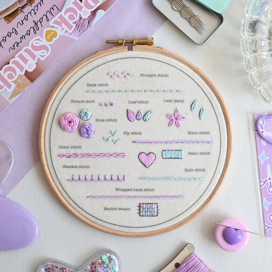 A wooden embroidery hoop displaying various stitch patterns and names on natural fabric, surrounded by craft supplies including a spool of lavender thread, a pin cushion, and a booklet on a pastel desk background.