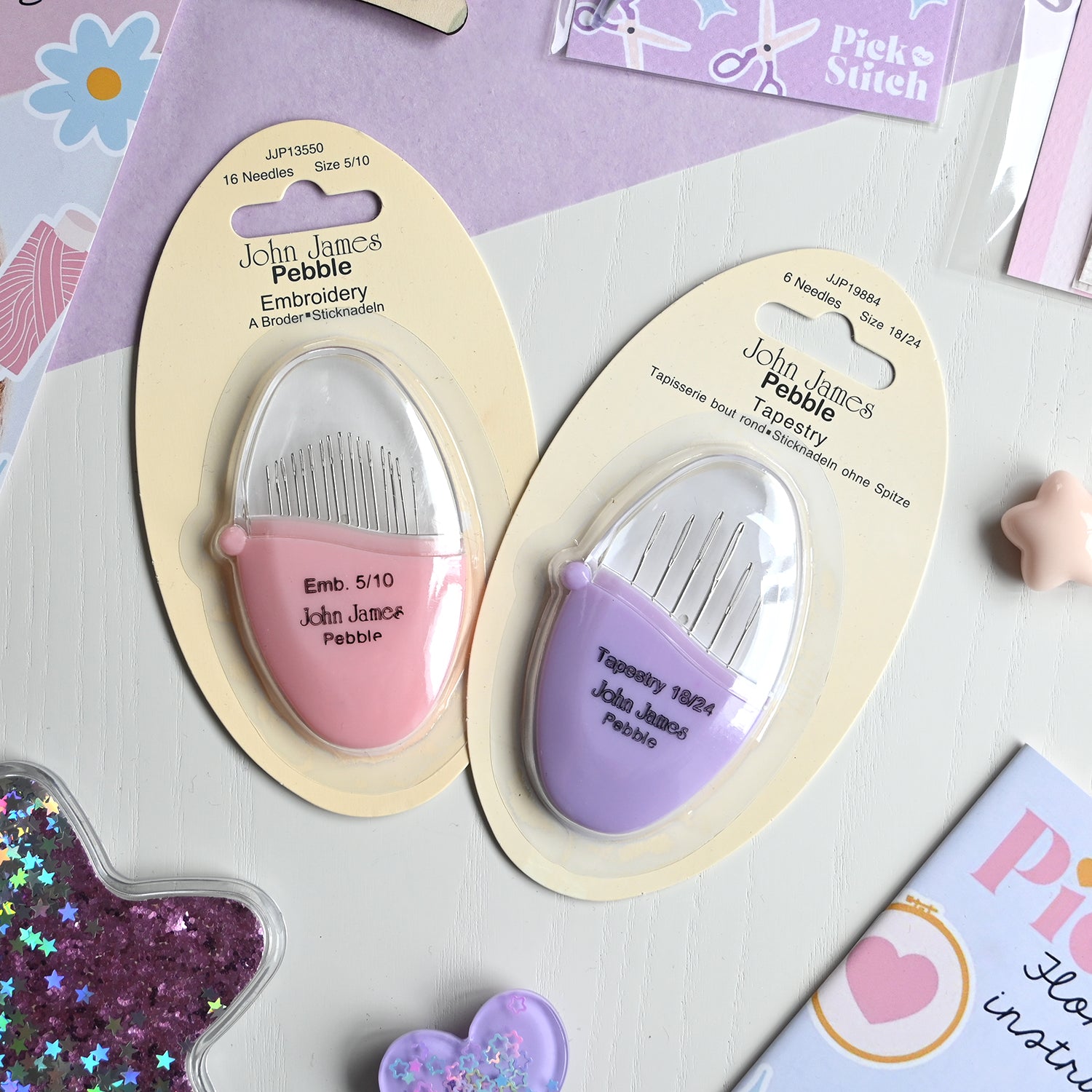 Two John James needle Pebble embroidery tools in packaging on a craft-themed surface.