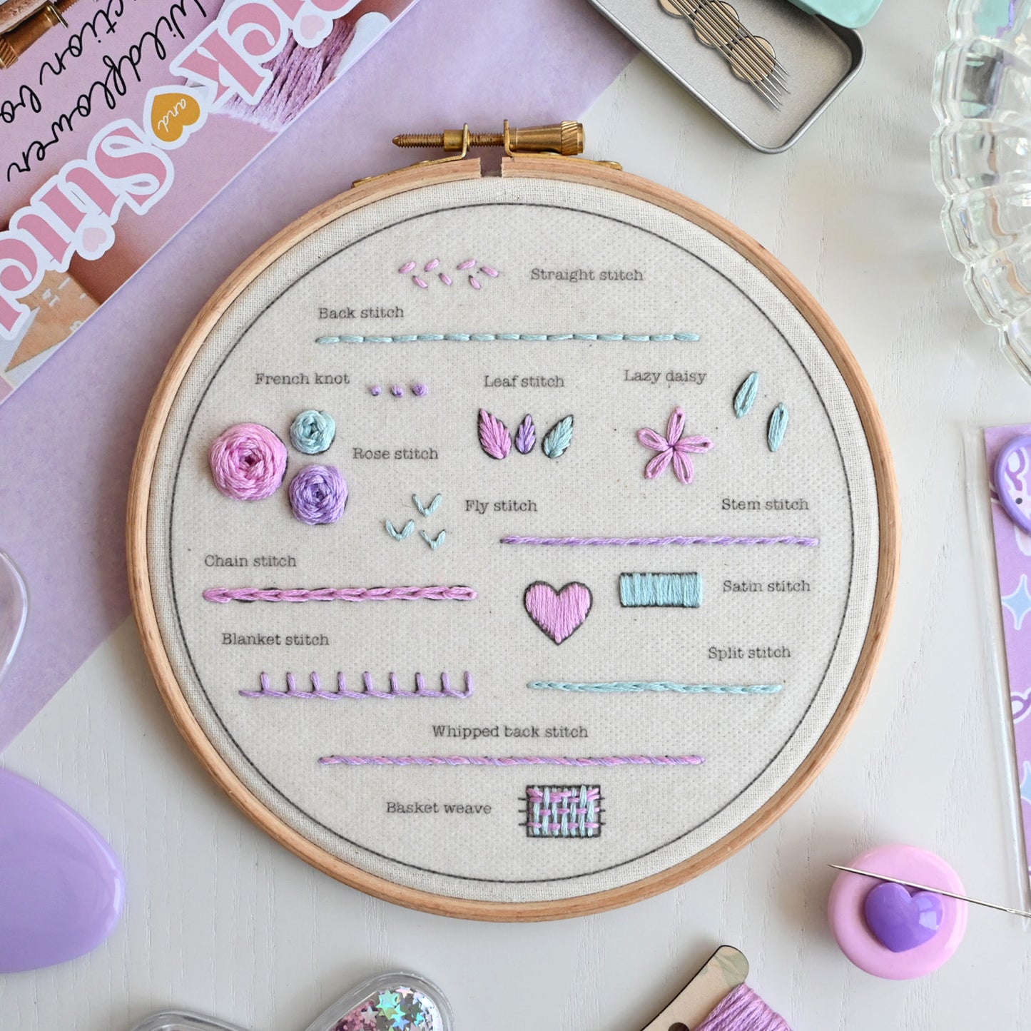 A wooden embroidery hoop displaying various stitch techniques and sample stitches on beige fabric, surrounded by colorful embroidery threads, a pink heart-shaped tool, and other crafting accessories on a light surface.