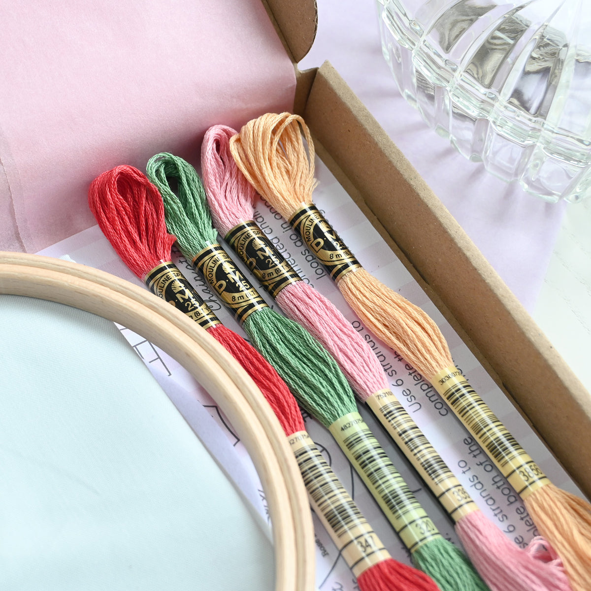 Four skeins of embroidery floss in coral red, sage green, light pink, and peach, arranged in a wooden hoop next to an open box of matching thread spools.