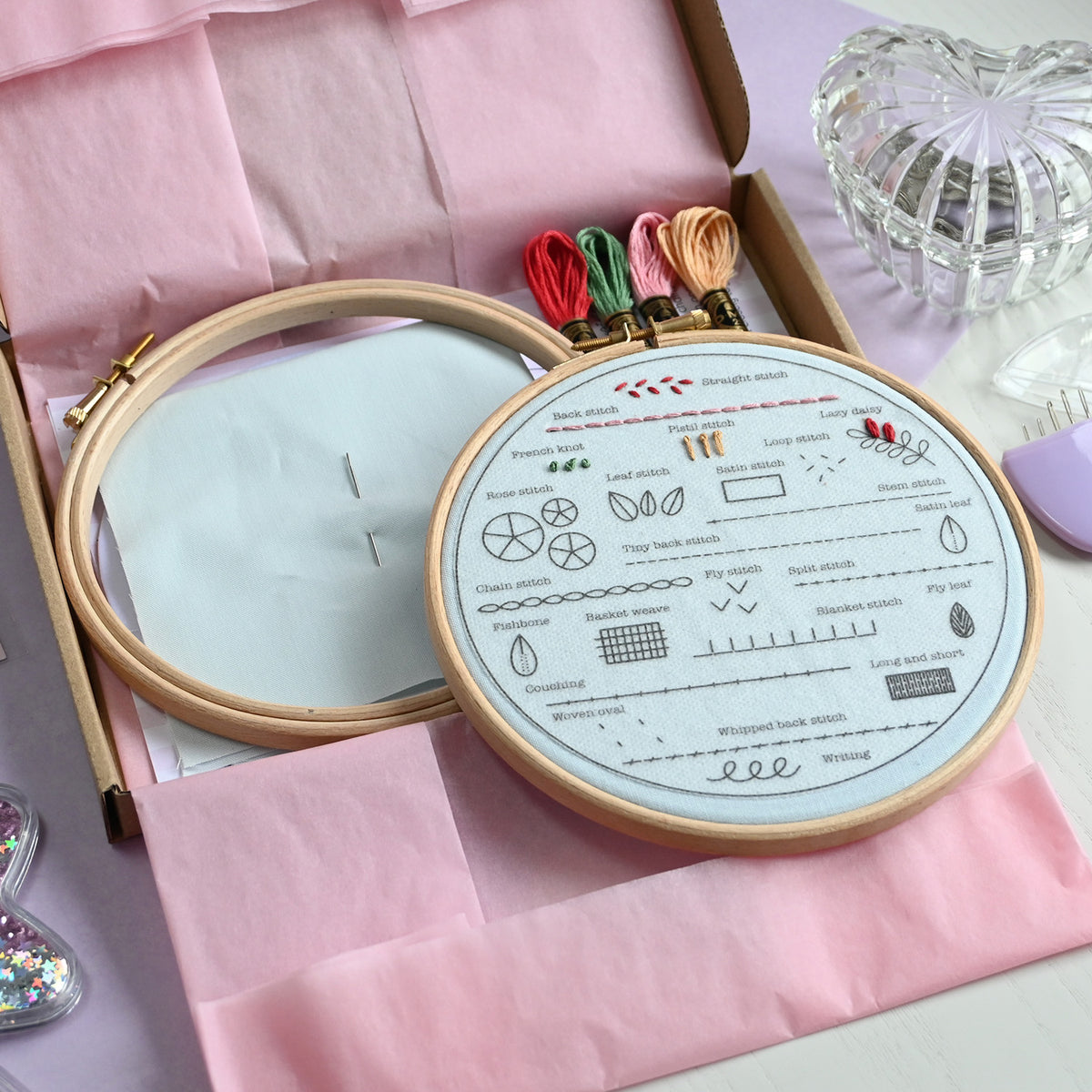 An embroidery kit displayed in an open box with pink tissue paper. The kit includes a wooden embroidery hoop with a printed stitch guide, white embroidery floss, colorful thread spools, and a glass heart-shaped dish on a lavender surface.