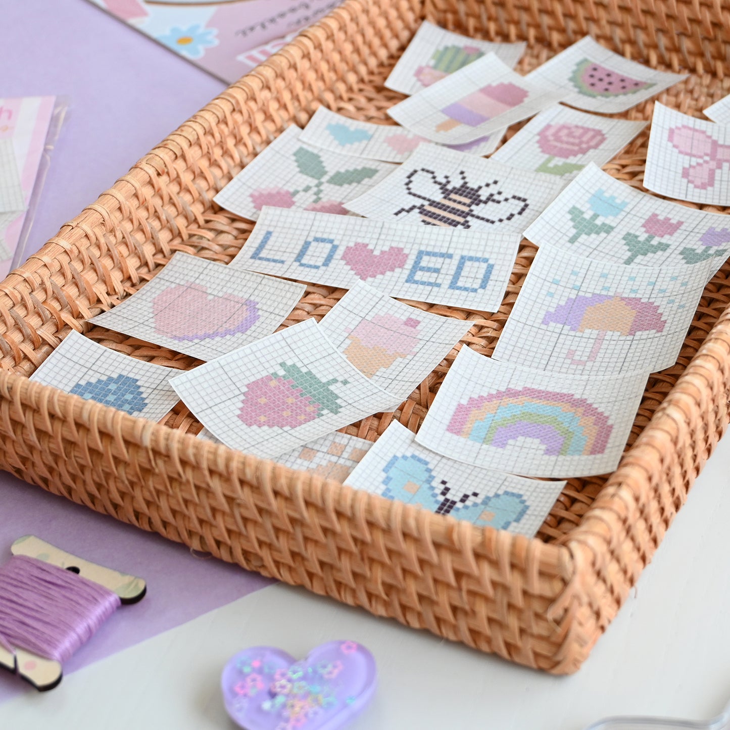 Cross-stitch patches on peel and stick paper with a wicker basket and embroidery tools on a light purple surface.